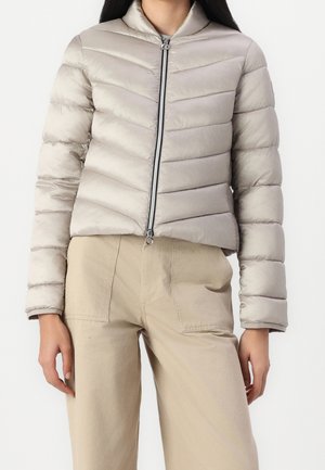 Femme portant une doudoune matelassée gris clair avec une fermeture éclair devant et un pantalon beige taille haute avec de grandes poches avant.