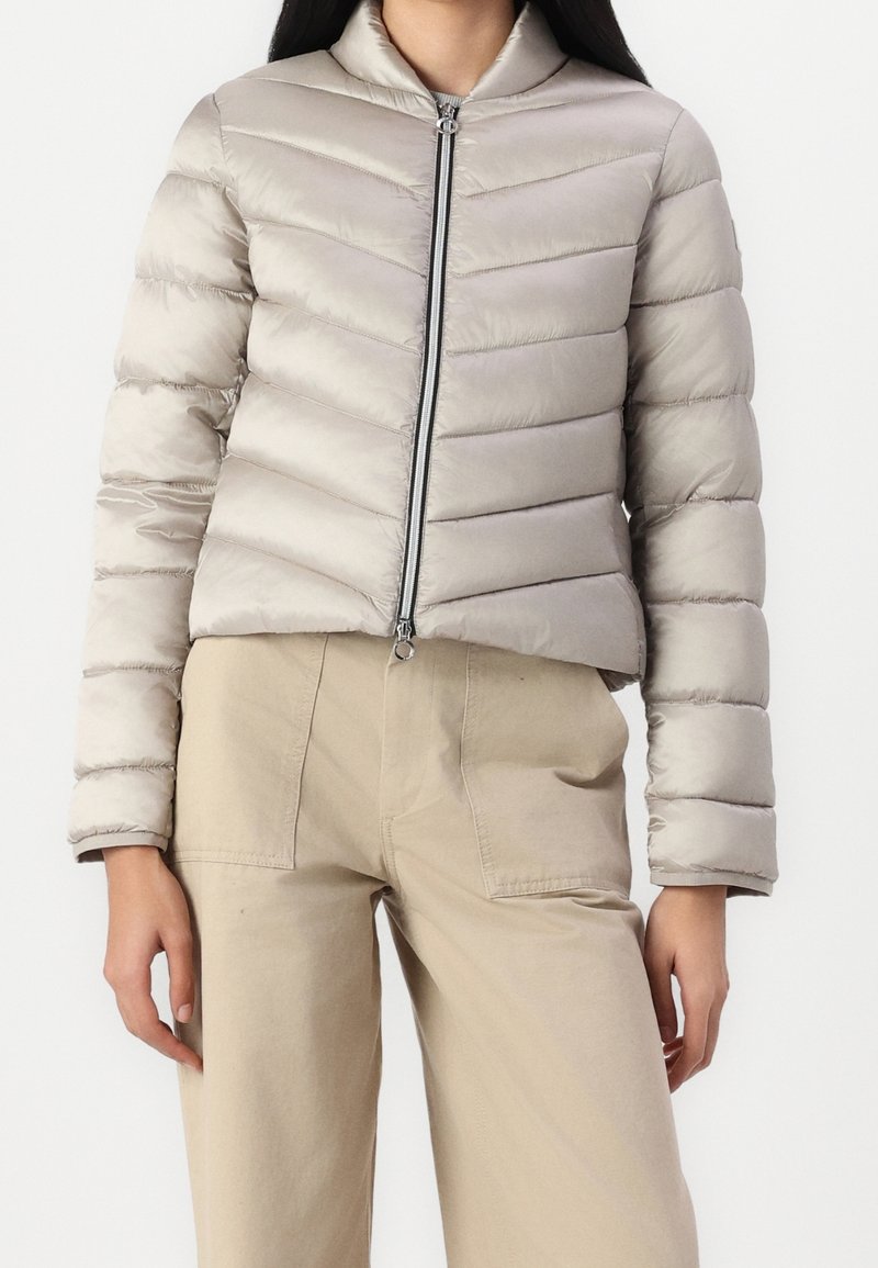 Femme portant une doudoune matelassée gris clair avec une fermeture éclair devant et un pantalon beige taille haute avec de grandes poches avant.