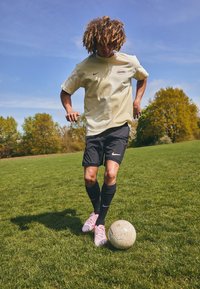 Athlète portant un t-shirt Nike jaune clair, un short noir et des crampons roses, en train de dribbler un ballon de football sur l'herbe sous un ciel bleu.