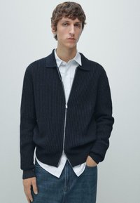 Cardigan blu navy a coste con colletto, zip frontale e polsini a contrasto. Indossato sopra una camicia bianca, abbinato a jeans blu. Design pulito e strutturato.