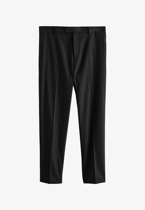 Mężczyzna ubrany w czarne eleganckie spodnie, czarne eleganckie buty, białą koszulę i szary krawat, stojący z jedną ręką w kieszeni na jednolitym tle.