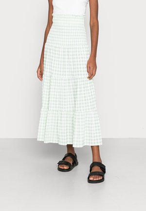 Vrouw draagt een lichtgroene en witte geruite gelaagde midi-rok, witte top en zwarte sandalen met open tenen, staand tegen een effen achtergrond.