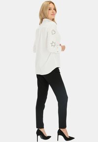 Biała koszula z długimi rękawami, ozdobiona dekoracyjnymi sercami i gwiazdkami z kryształków na rękawach. Zestawiona z czarnymi dopasowanymi spodniami i czarnymi szpilkami.