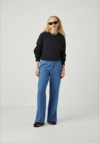 Donna con capelli biondi ricci, che indossa occhiali da sole neri, un felpa nera, jeans a gamba larga blu e sneaker nere con accenti arancioni.