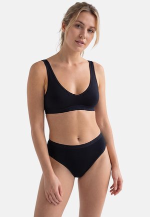 Jeune femme aux cheveux blonds portant un bralette noir et un slip taille haute assorti, debout devant un fond clair uni.