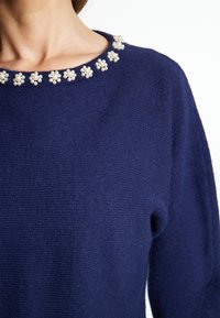 Marineblauer Wollpullover mit Rundhalsausschnitt, der mit perlenähnlichen floralen Verzierungen geschmückt ist. Weiche Textur, etwas lockere Passform.
