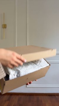 Brown cardboard box being opened, revealing crinkled white tissue paper with black diagonal stripes inside. Simple, rectangular design.