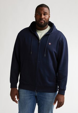 Marineblauwe hoodie met ritssluiting, trekkoorden, kangoeroezakken en logopatch. Draagt over een lichtgroen shirt. Soepele textuur, casual pasvorm.