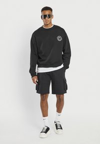 Homme portant un sweat-shirt noir avec un logo smiley, un short cargo noir, des chaussettes blanches, des baskets noires et blanches, et des lunettes de soleil noires surdimensionnées, debout.