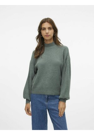 Groene gebreide trui met een hoge hals, verlaagde schouders en geplooide lange mouwen, gecombineerd met blauwe denim jeans. Glad textuur.
