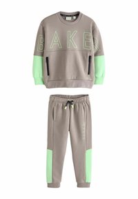 Zweiteiliger Trainingsanzug in Beige mit grünen Akzenten. Der Pullover trägt den "BAKER"-Druck und hat mit Reißverschlüssen versehene Taschen; die Hose hat ähnliche grüne Seitenpaneele.