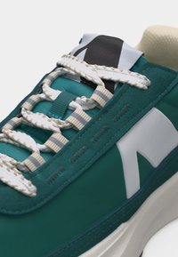 Pantof sport teal cu șireturi groase albe, cusături bej, guler căptușit și un logo mare alb pe lateral, deasupra unei tălpi albe groase.