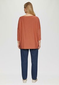 Roest-oranje losse top met een horizontaal textuurpatroon, driekwart mouwen, gecombineerd met donkerblauwe recht gesneden jeans en crèmekleurige schoenen.