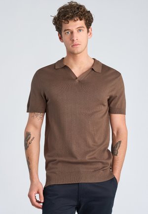 Młody mężczyzna o kręconych włosach, ubrany w brązową, krótki rękaw, dzianą koszulkę polo i ciemne spodnie, stoi z jedną ręką w kieszeni.