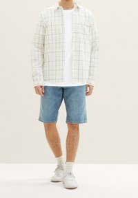 Camisa de cuadros de manga larga en blanco y verde sobre una camiseta blanca, combinada con pantalones cortos de mezclilla azul y zapatillas blancas con calcetines beige.