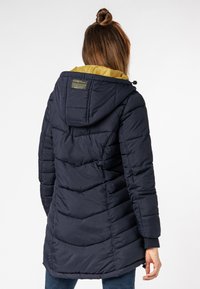 Vrouw met bruin haar in een knot, gekleed in een donkerblauwe gewatteerde jas met een capuchon die aan de binnenkant geel is, staande met haar rug naar de camera.