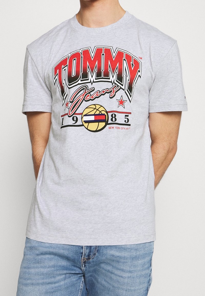 Mies, joka käyttää vaaleanharmaata T-paitaa, jossa on punainen ja musta "TOMMY Jeans 1985" logo, keltainen koripallograafikka ja farkut.