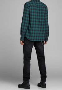 Camisa de franela a cuadros en verde y negro con puños abotonados, combinada con jeans negros que tienen bolsillos traseros y un acento de cadena, usados con botas negras.
