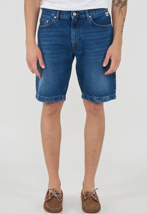 Uomo che indossa pantaloncini di jeans blu di media lunghezza, scarpe da barca in pelle marrone e una maglietta bianca, con tatuaggio visibile sul braccio sinistro.