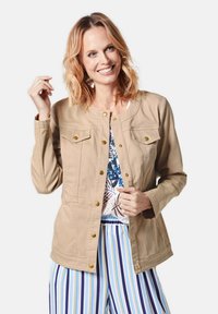 Beige casual jasje met gouden drukknopen, twee borstzakken en een ronde halslijn. Draagt over een blauwgepatenteerde top en gestreepte broek.