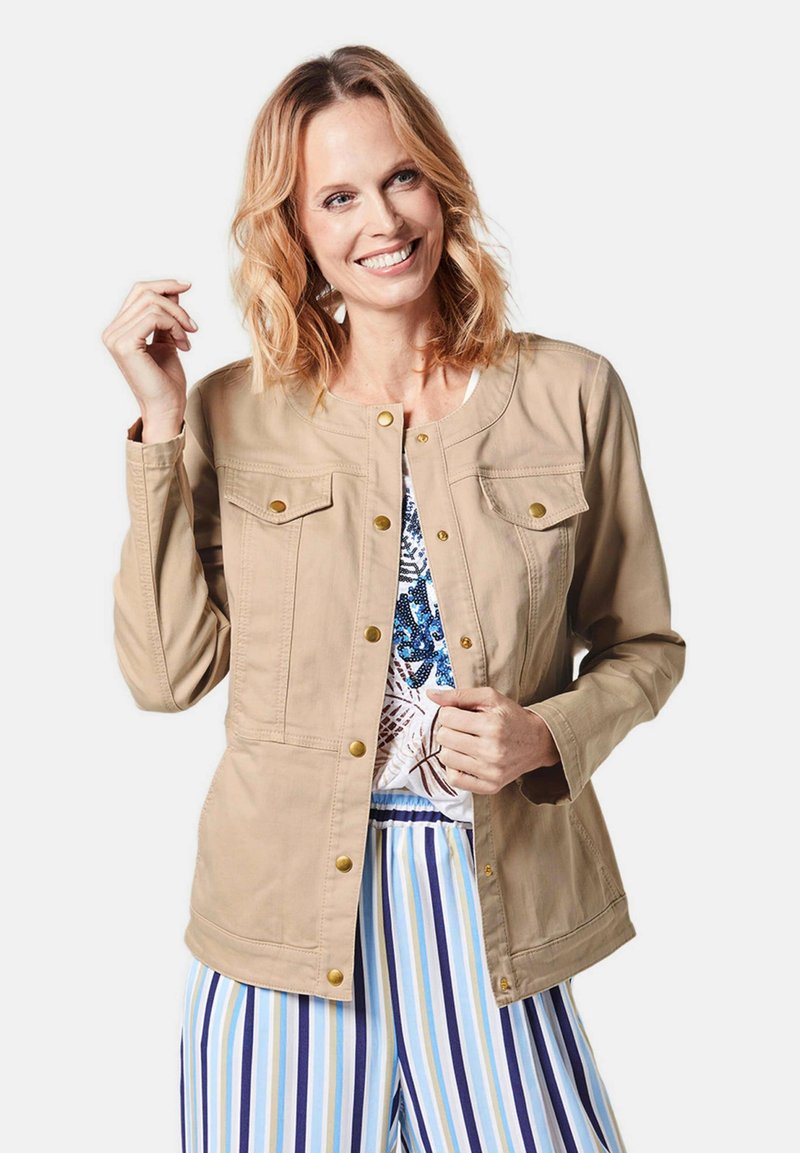 Beige casual jasje met gouden drukknopen, twee borstzakken en een ronde halslijn. Draagt over een blauwgepatenteerde top en gestreepte broek.