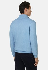 Pull bleu clair avec un col haut, texture tricotée, et poignets et ourlet côtelés. Associé à un pantalon en velours côtelé noir. Vue de dos et de côté visible.