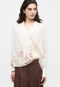 Γυναίκα που φοράει λευκό πουκάμισο με εκτυπώσεις κοχυλιών και αστερίας, συνδυασμένο με καφέ παντελόνια ευρέος ποδός, κοιτάζοντας προς το πλάι.