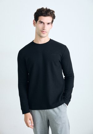 Schwarzes Langarmshirt mit strukturiertem Muster, klassischem Rundhalsausschnitt und einem dezenten grauen Logo. Getragen mit grauen Jogginghosen.