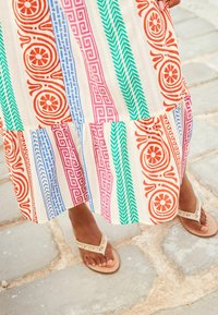 Robe longue multicolore aux motifs floraux et géométriques en rouge, bleu, rose et vert. Sandales beiges avec des accents décoratifs.
