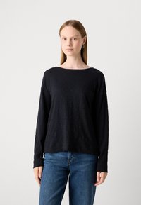 Young woman with straight blonde hair wearing silver hoop earrings, a loose black long-sleeve top with pocket, and blue jeans, standing.
