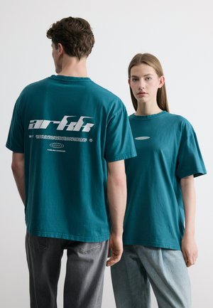 Uomo e donna indossano t-shirt oversize color teal; l'uomo è di spalle mostrando un logo bianco sul retro, la donna è di fronte con un piccolo logo sul davanti.