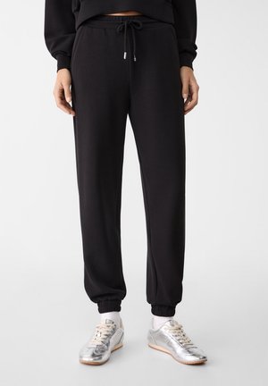 Person wearing black drawstring sweatpants with elastic cuffs and silver sneakers, standing on a white background.