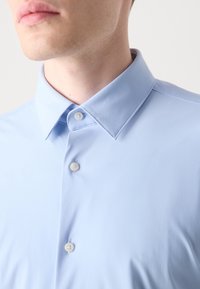 Homme portant une chemise bleu clair avec un col pointu et des boutons blancs, visible du cou jusqu'à la partie supérieure de la poitrine.