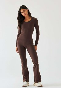 Brązowa dopasowana bluzka z długim rękawem w połączeniu z pasującymi, rozkloszowanymi legginsami wykonanymi z elastycznego materiału o gładkiej fakturze. Całość uzupełniają białe tenisówki.