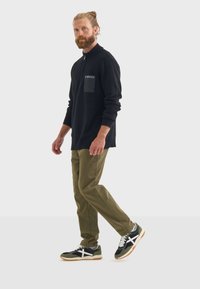 Schwarzer Reißverschluss-Pullover mit strukturierter Oberfläche und einer Brusttasche, kombiniert mit olivgrünen Hosen und schwarzen Sneakers mit weißen Akzenten.