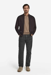 Mörklila stickad cardigan, ljusbrun polo skjorta, grå byxor och beige sneakers. Modellen står med händerna i fickorna mot en vit bakgrund.