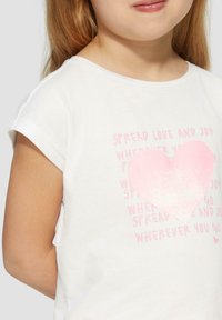 Witte katoenen t-shirt met korte mouwen, voorzien van een grote roze hartafbeelding en tekst die luidt "Verspreid liefde en vreugde" in lichtroze.