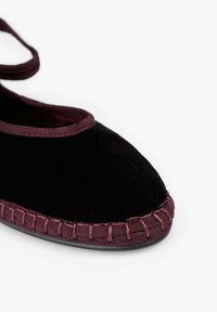 Zapato de terciopelo negro con puntera redondeada, ribete burdeos y costuras decorativas a lo largo de la suela. Correa para un ajuste seguro.