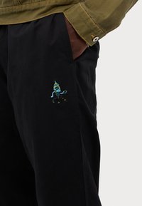 Black trousers feature a playful, colorful embroidered design of a character on the left pocket. Fabric is smooth with a slight sheen.