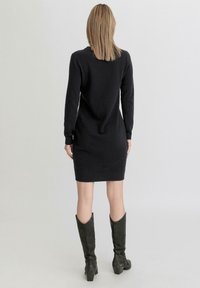 Abito nero a maglia con maniche lunghe, forma aderente, polsini a coste e una texture liscia. Abbinato a stivali scuri a ginocchio.