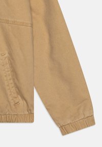 Manica di giacca leggera beige con una texture liscia, polsini elasticizzati e una tasca laterale con dettaglio cucito.