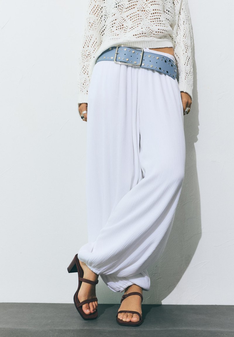 Personne portant un pantalon blanc ample avec une large ceinture bleue cloutée, un pull en tricot blanc, des bagues et des sandales marron à talons, debout contre un mur.