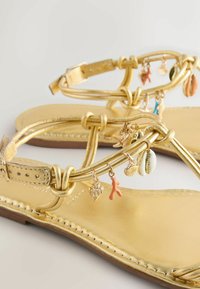 Sandales plates en or avec des brides à la cheville ornées de petits charms en forme de coquillages et de corail sur un fond clair.