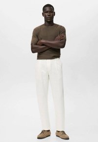 Chemise marron à rayures sur les manches courtes, pantalon blanc ajusté et chaussures à lacets marron. Le modèle se tient les bras croisés devant un fond neutre.