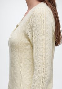 Femme aux longs cheveux bruns portant un cardigan tricoté de couleur crème avec un motif géométrique subtil et une fermeture par boutons au niveau de l'encolure.