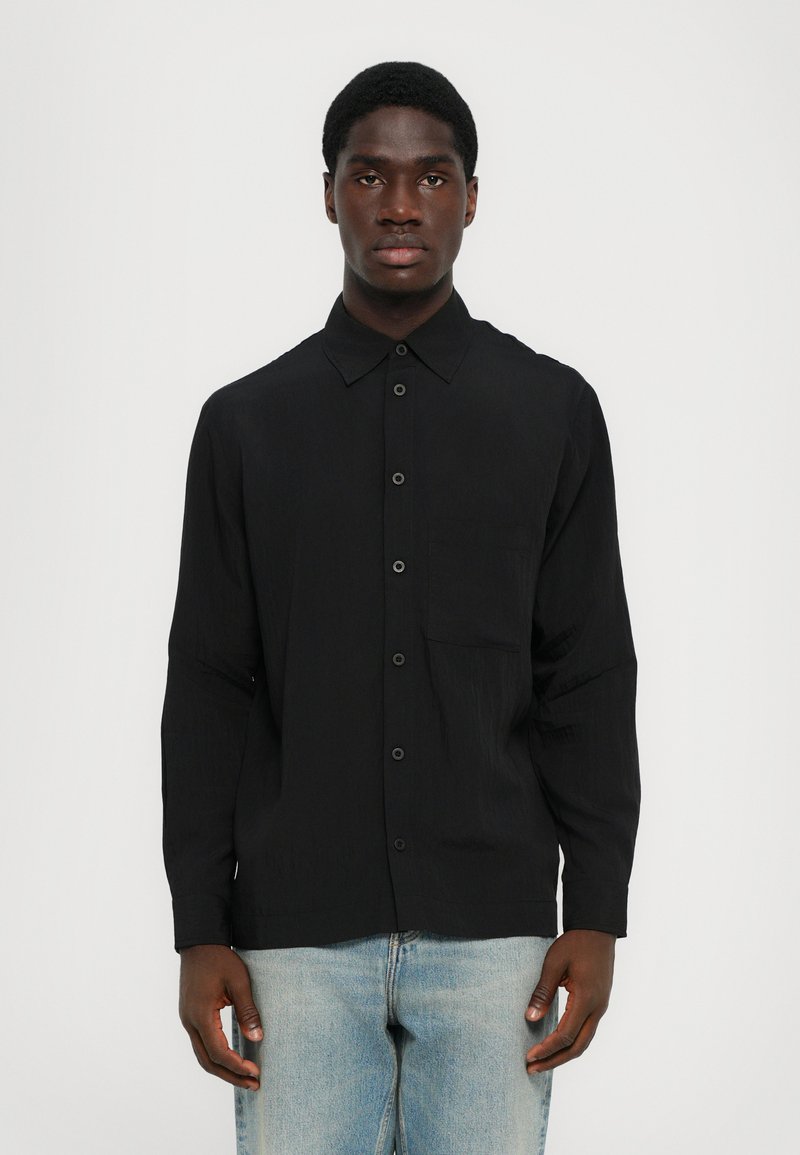 Mustast pikast käistest särgist, millel on nööpidega krae, eesmine tasku ja sile tekstuur, koos helesiniste teksadega.