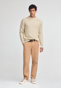 Pull en maille côtelée beige, chino beige, et baskets blanches. Design simple, texture douce, et coupe décontractée. Détail de marque minimal sur le pull.