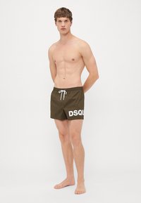 Junger Mann steht barfuß in braunen Badeshorts mit weißem DSQUARED2-Logo und weißem Kordelzug vor einem schlichten weißen Hintergrund.