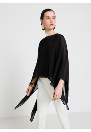 Frau trägt eine durchsichtige schwarze asymmetrische Bluse, weiße Hose, goldene Blätterohrringe und Armbänder, steht vor einem schlichten hellen Hintergrund.