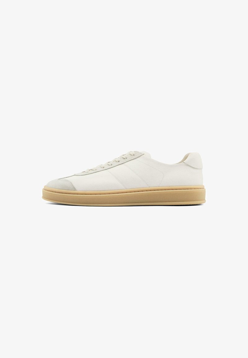 White suede sneakers with a smooth texture, round toe, lace-up front, and a tan rubber sole. Features subtle side stripes and heel accents.